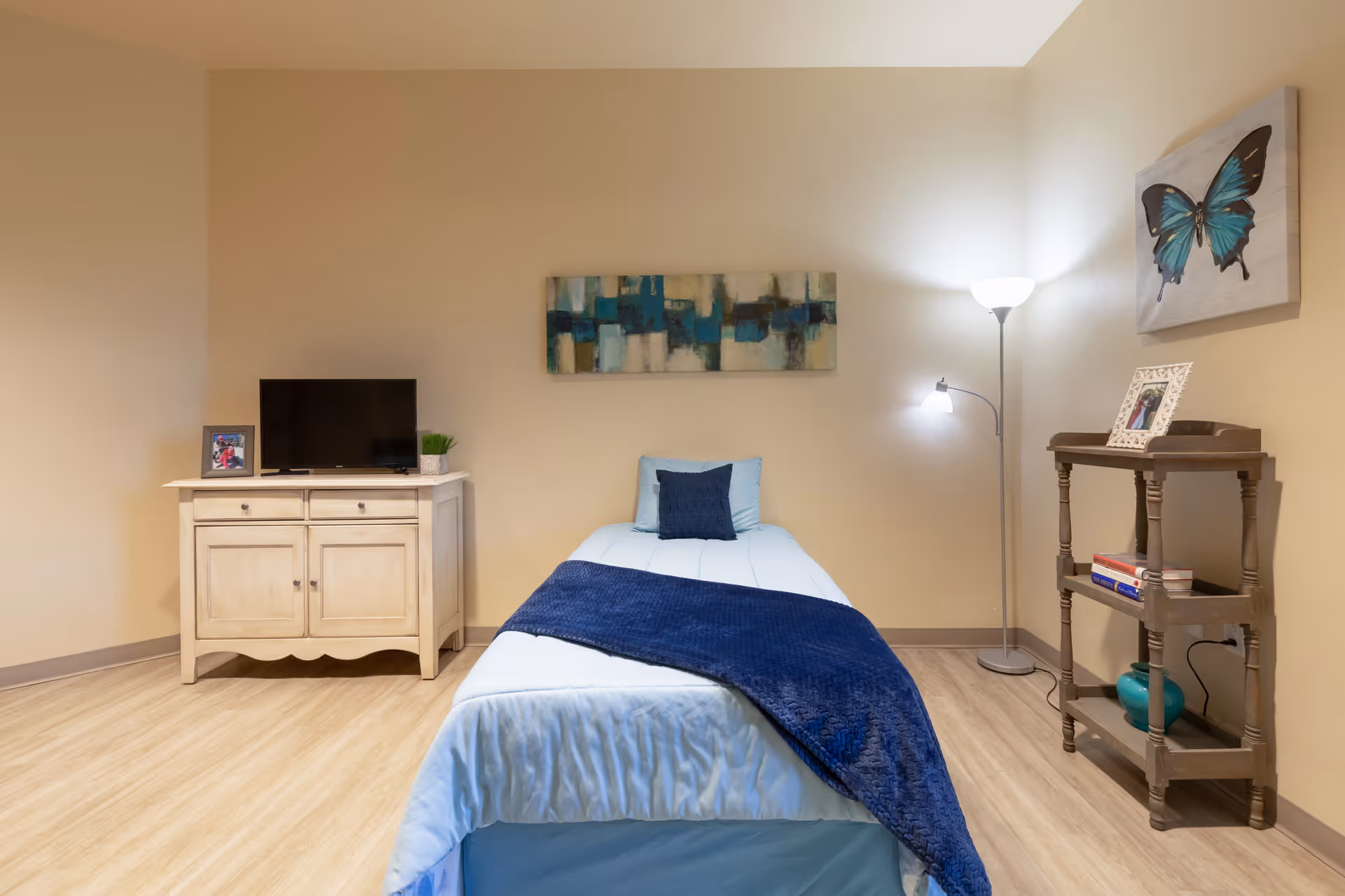 A small, neatly arranged bedroom with a single bed covered in light blue bedding and a dark blue throw blanket. Above the bed is a rectangular abstract painting with blue and beige tones. To the left of the bed is a cream-colored cabinet with a small flat-screen TV, a framed photo, and a small plant. To the right of the bed is a wooden shelf holding books, a framed photo, and a turquoise vase. A floor lamp with two lights stands next to the shelf. The room has beige walls and light wood flooring.