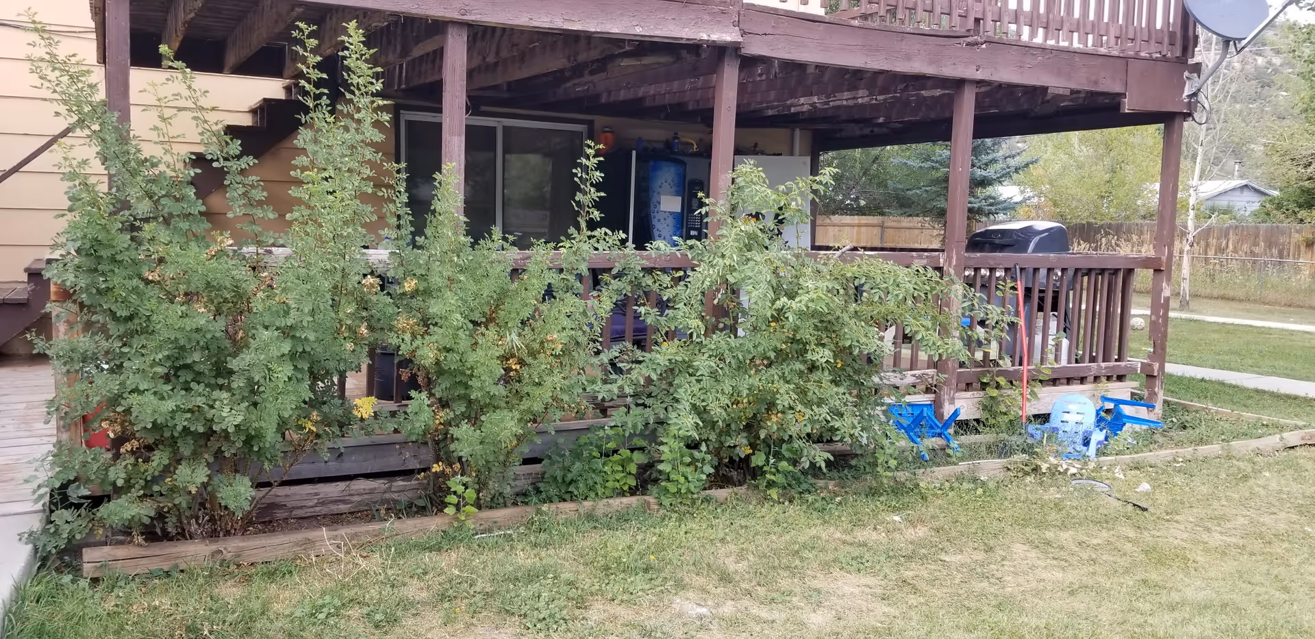 Outdoor view of a wooden deck attached to a building with green bushes in front. The deck has a railing and a staircase on the left side. There is a vending machine and a grill on the deck, with some blue plastic chairs overturned on the grass nearby. Trees and a wooden fence are visible in the background.