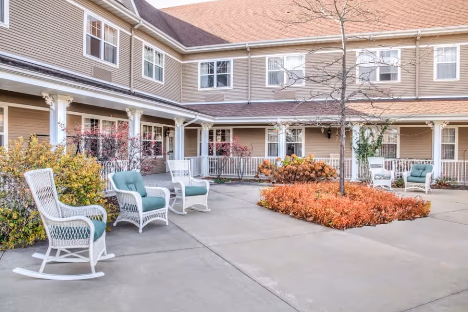 Courtyard with white wicker chairs and green cushions around a planted island in front of a two-story senior living building.
