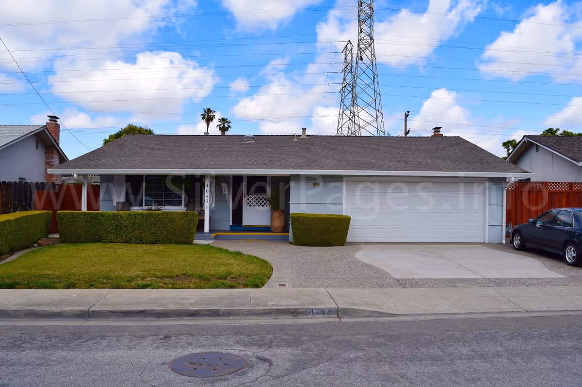 Single-story suburban house front with a two-car garage, trimmed hedges, driveway and a parked car under a partly cloudy sky.