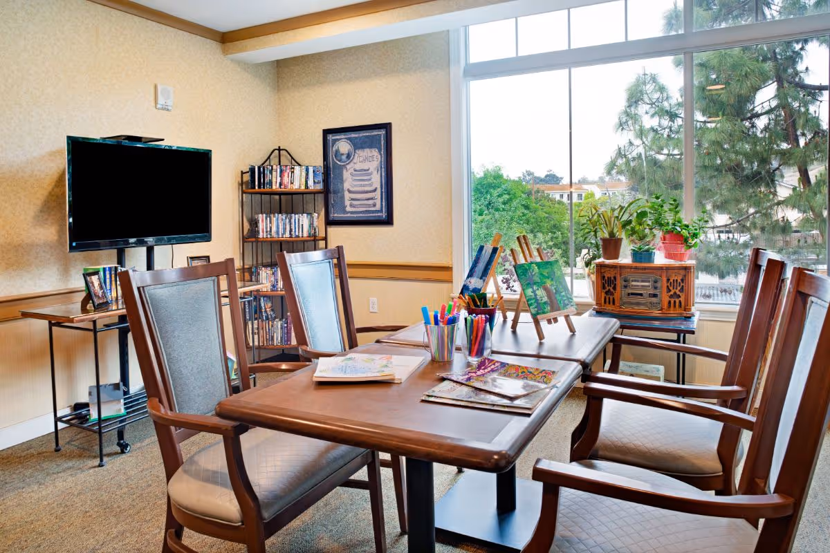 Sunlit activity room with a wooden table and chairs set up with art supplies and small easels, a TV and bookshelf, and a large window overlooking trees.