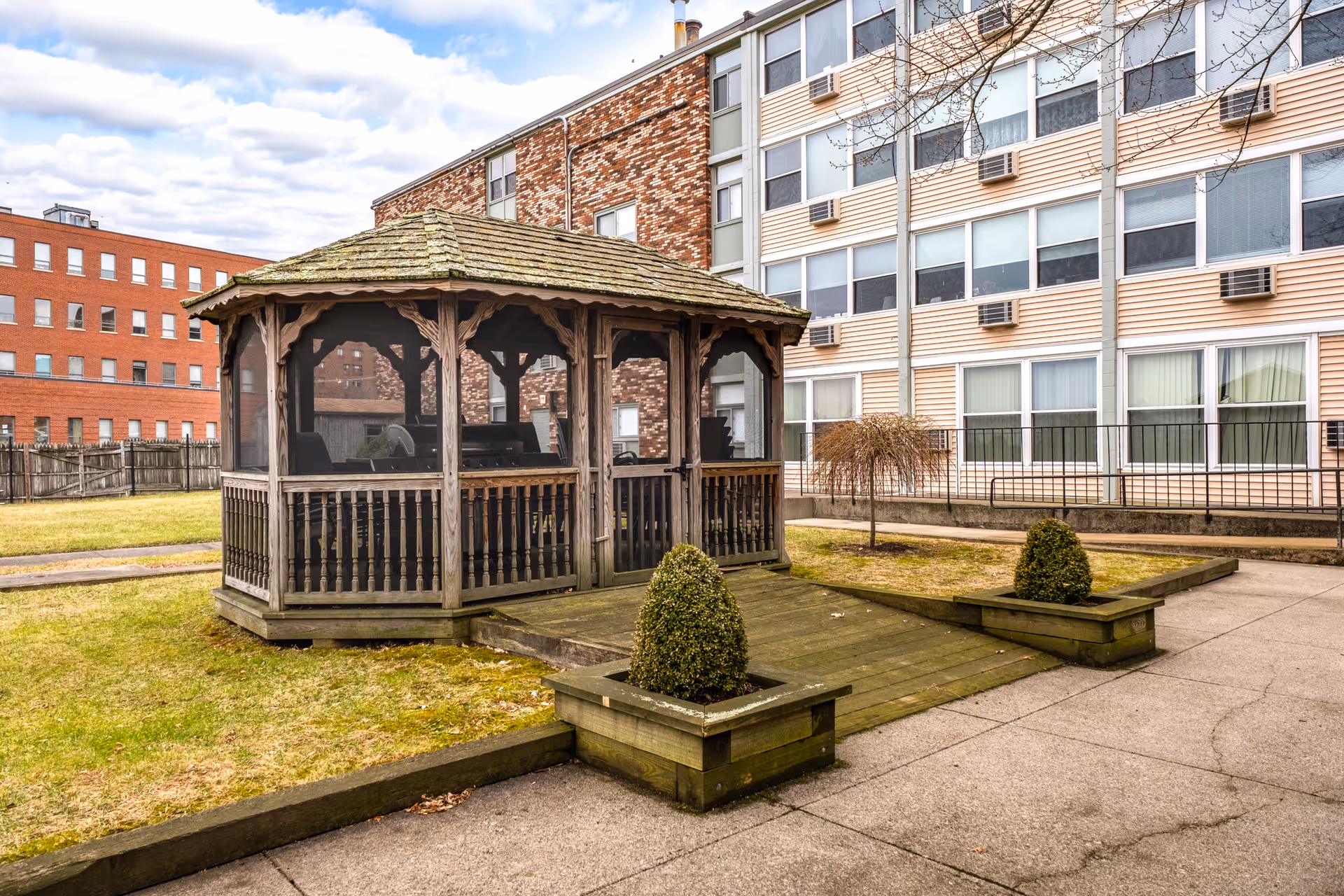 Outdoor area of a senior living facility featuring a wooden gazebo with a shingled roof and screened sides, surrounded by a grassy lawn and paved walkways. The background shows a multi-story building with numerous windows and air conditioning units.
