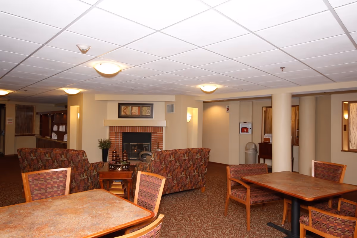 A senior living facility common area with patterned armchairs and wooden tables with chairs. There is a brick fireplace with a decorative piece above it, and the room is softly lit with ceiling lights. The walls are beige, and the carpet has a patterned design.