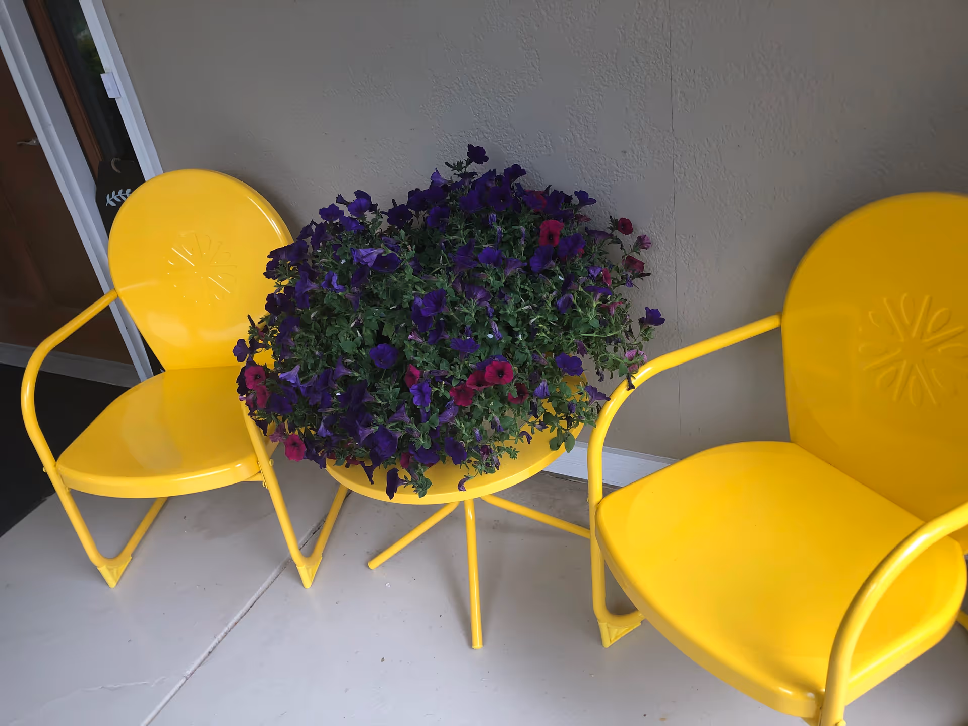 Two yellow metal chairs and a small yellow table holding a planter of purple and magenta flowers on a porch.