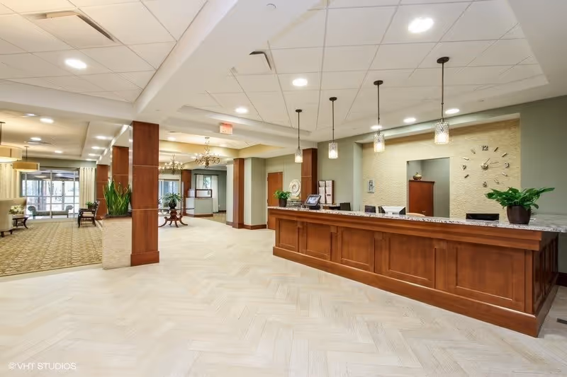 A spacious reception area in a senior living facility with a long wooden front desk, pendant lights hanging from the ceiling, potted plants on the desk, and a wall clock behind the desk. The area has light-colored flooring and a carpeted seating area with chairs and tables visible in the background near large windows.