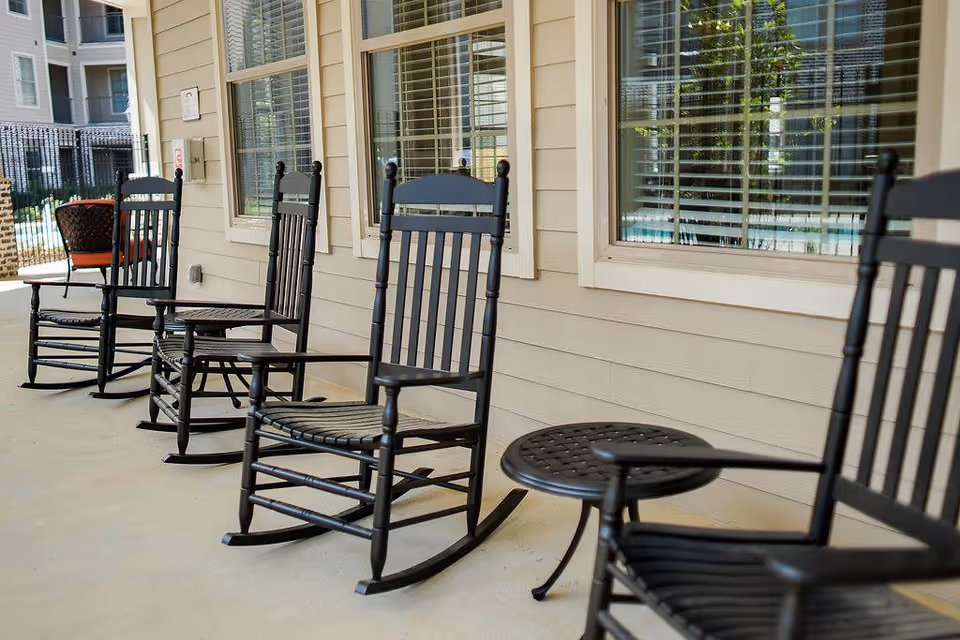 A row of black wooden rocking chairs and a small round table on a covered porch outside a building with beige siding and multiple windows with white blinds.