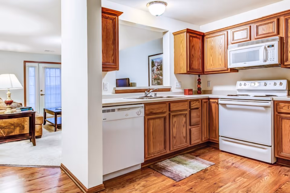 A bright kitchen with wooden cabinets, white appliances including a stove, microwave, and dishwasher. The kitchen has a hardwood floor and a small rug in front of the sink. Adjacent to the kitchen is a living room area with a lamp on a wooden side table, a sofa, a coffee table, and French doors with sheer curtains.