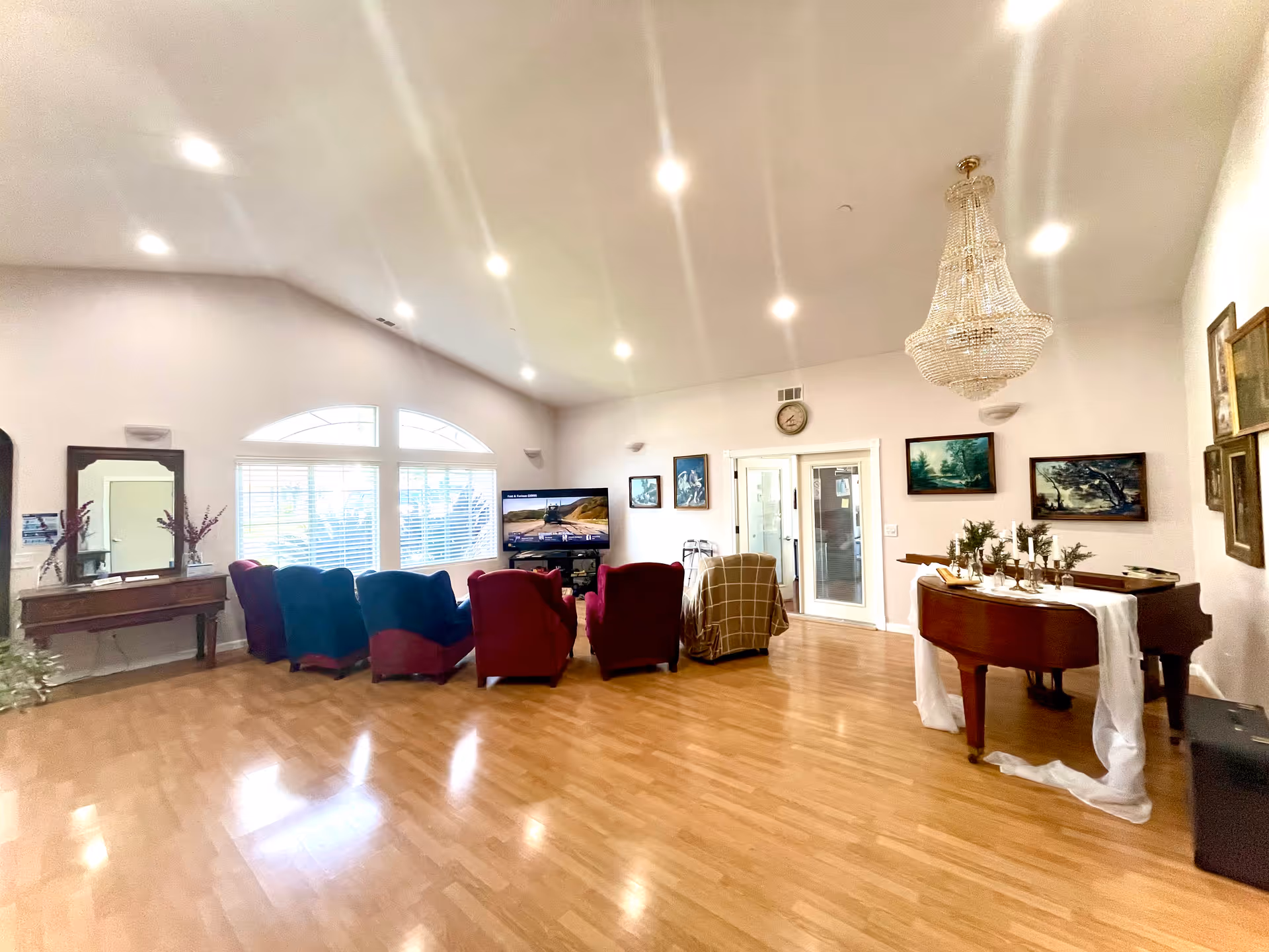 Spacious senior living common room with hardwood floors, a row of upholstered chairs facing a television, a grand piano under a chandelier, and large arched windows.