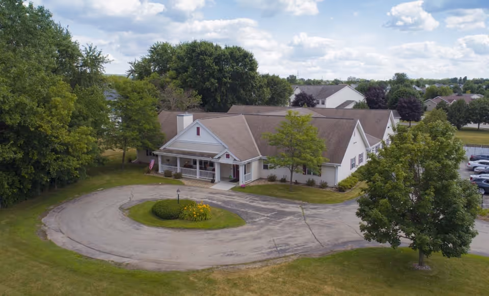 Aerial view of a single-story assisted living facility building with a circular driveway and landscaped greenery, surrounded by trees and a parking area under a partly cloudy sky.