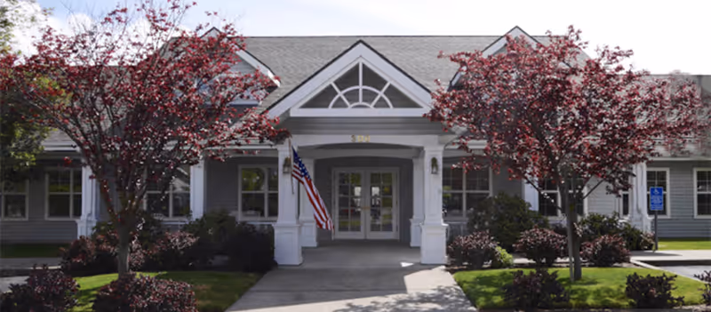 Front view of Trustwell Living at Suncrest Place entrance with American flag and trees.