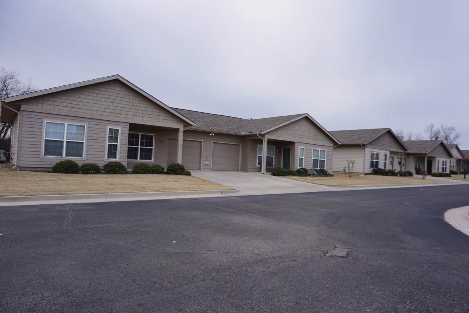Single-story beige senior living building with attached units and garages facing a paved road under an overcast sky.