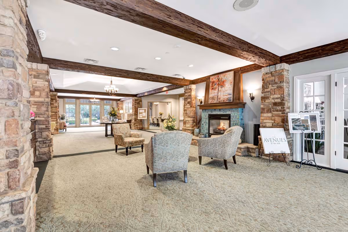A spacious and well-lit living room area in a senior living facility featuring stone pillars, wooden ceiling beams, a fireplace with a colorful painting above it, three patterned armchairs arranged around the fireplace, and a table with flowers in the background. Large windows and glass doors allow natural light to fill the room.