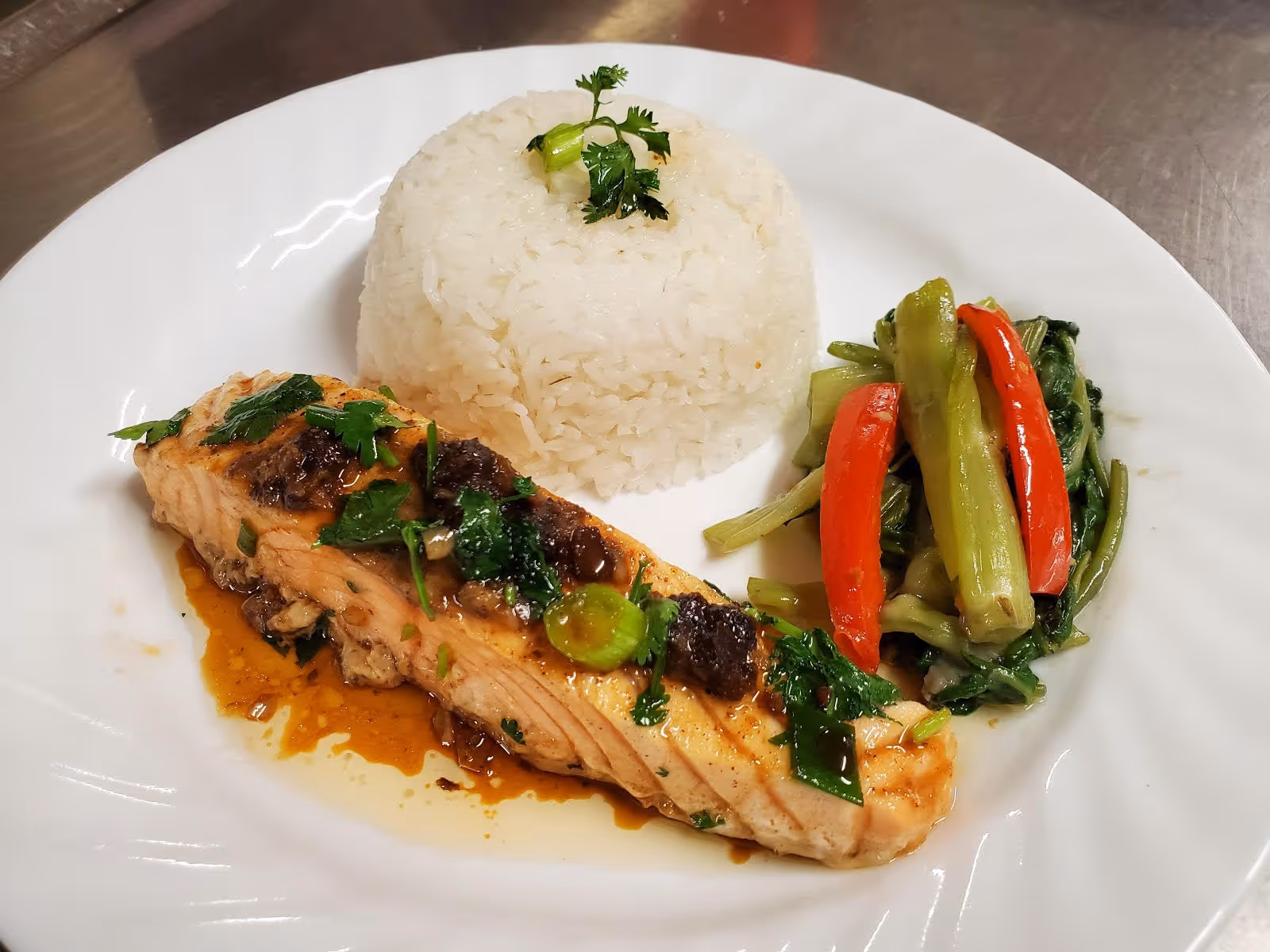A plated meal consisting of a piece of cooked salmon garnished with herbs and sauce, a serving of white rice topped with a small sprig of parsley, and a side of cooked mixed vegetables including red bell peppers and green stalks.