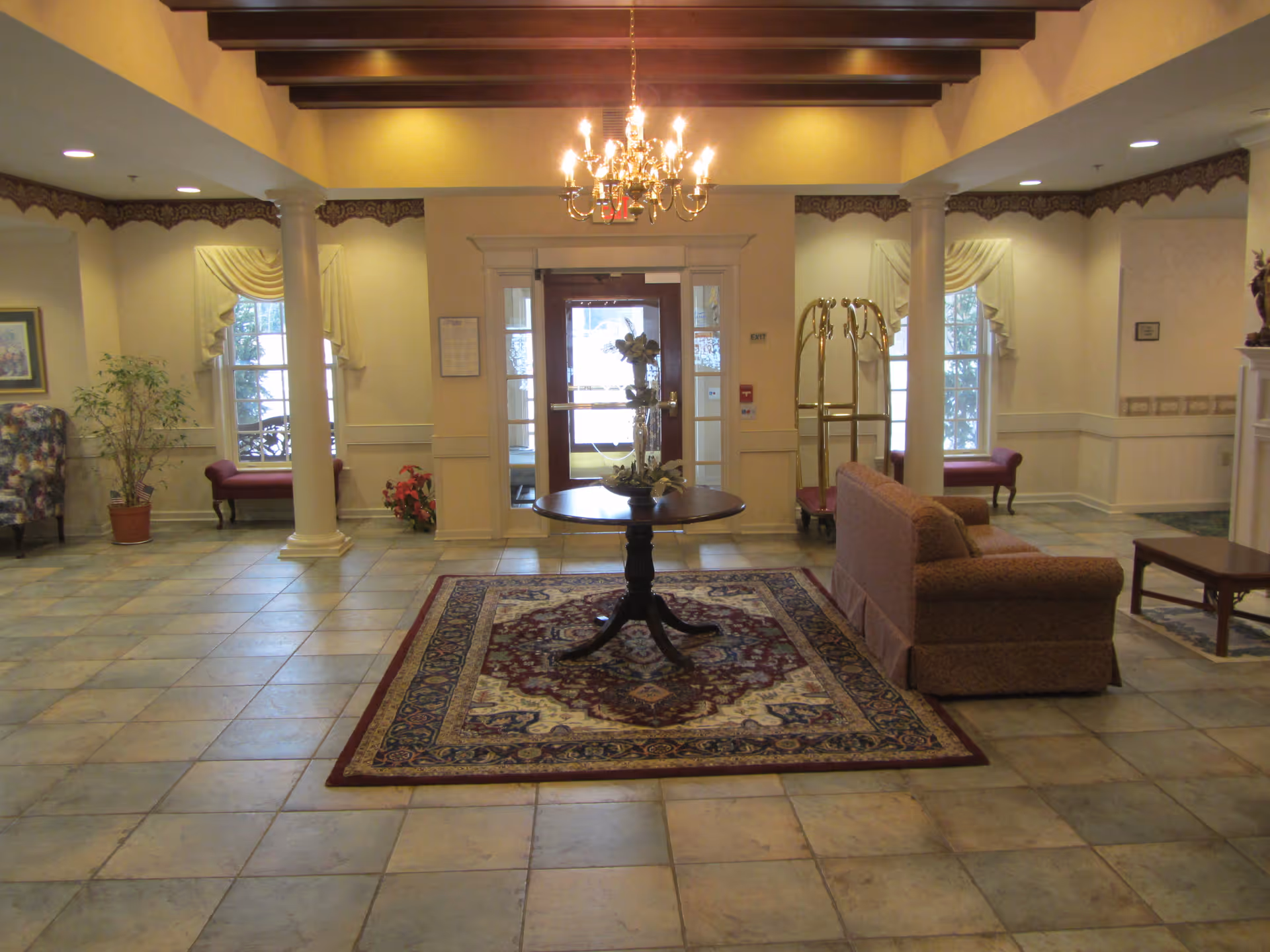 Spacious assisted living lobby with tiled floor, a central round table on an area rug, seating, chandelier, and entry doors.