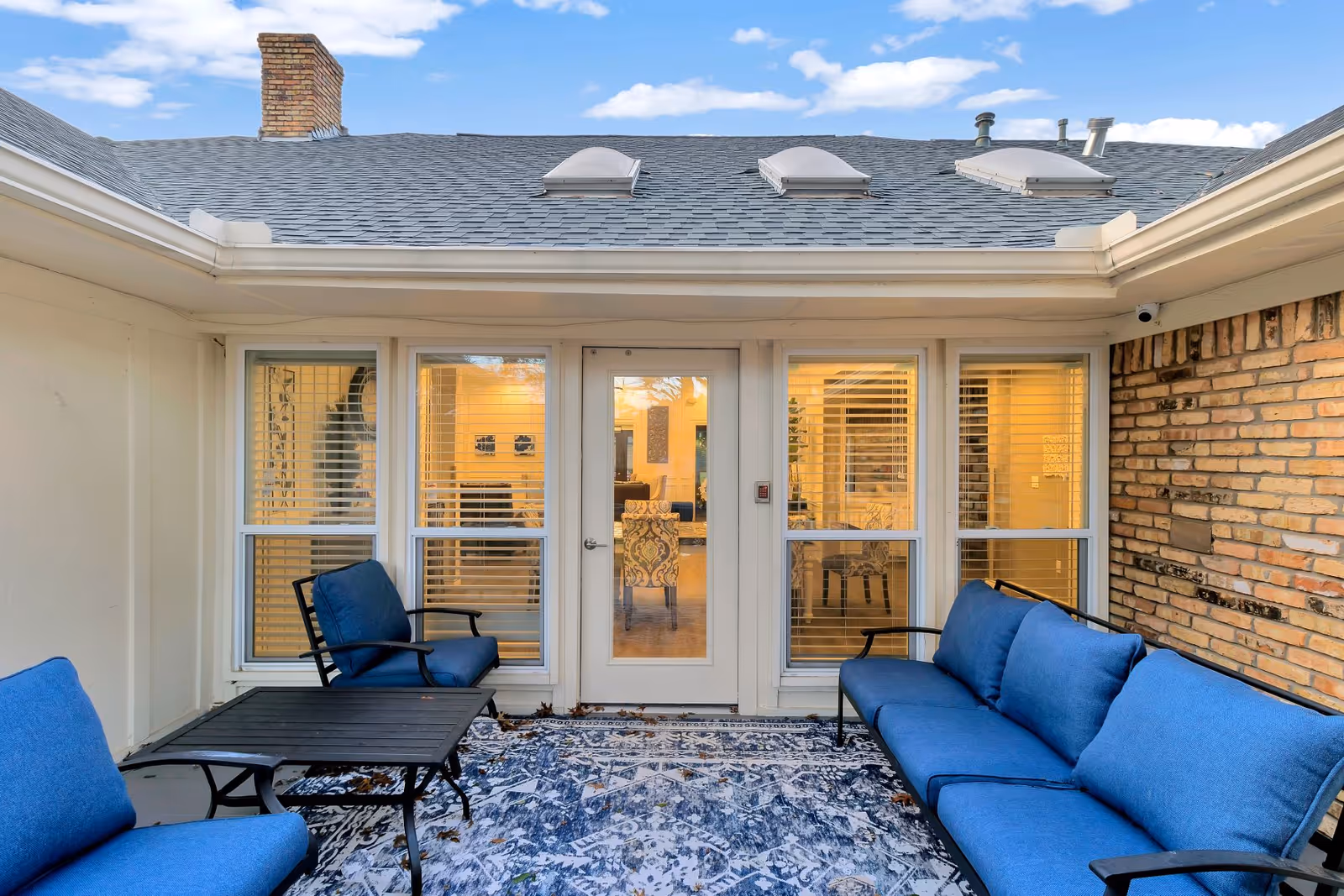 Outdoor patio area with blue cushioned seating including a sofa and chairs, a black coffee table, and a patterned outdoor rug. The patio is adjacent to a building with large windows and a glass door leading inside, showing a warmly lit interior.