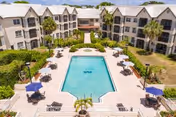View of an outdoor swimming pool surrounded by lounge chairs and umbrellas, situated in the courtyard of a multi-story residential building with balconies and palm trees.