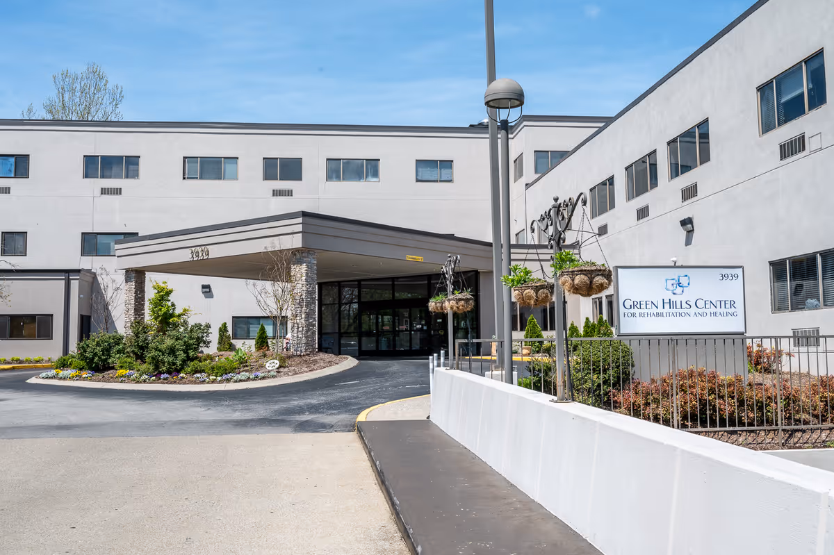 Exterior view of Green Hills Center for Rehabilitation and Healing, showing a multi-story building with a covered entrance, landscaped bushes and flowers, hanging plants, and a sign with the facility name.