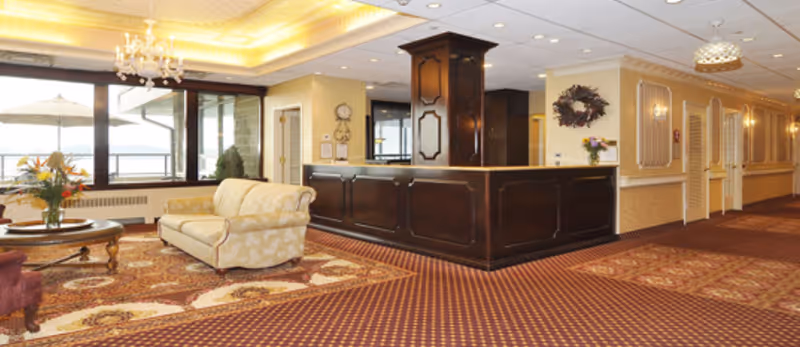 A spacious and warmly lit interior area featuring a dark wooden reception desk with a column in the center. To the left, there is a seating area with a cream-colored sofa and a round wooden coffee table with a flower arrangement. Large windows behind the seating area allow natural light to enter. The floor is covered with patterned carpets in red and beige tones. The walls are decorated with a wreath and sconces, and there is a hallway leading to other rooms on the right side.