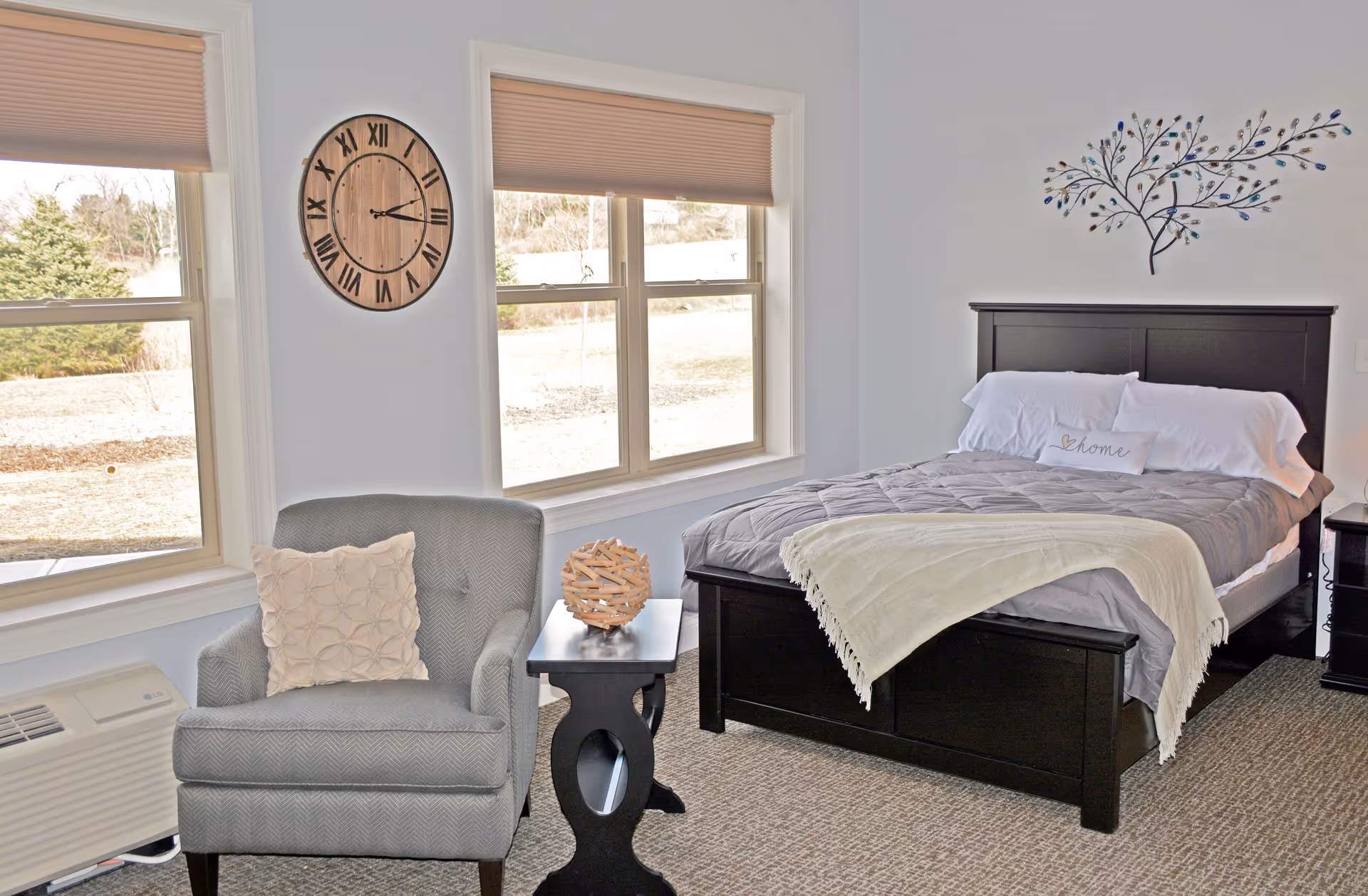 A bright bedroom with a large window showing an outdoor view. The room features a black wooden bed with gray bedding and a white throw blanket, a decorative pillow with the word 'home', a gray armchair with a textured cushion, a small black side table with a decorative wooden orb, a wall clock with Roman numerals, and a metal wall art piece shaped like a tree with blue and green leaves.
