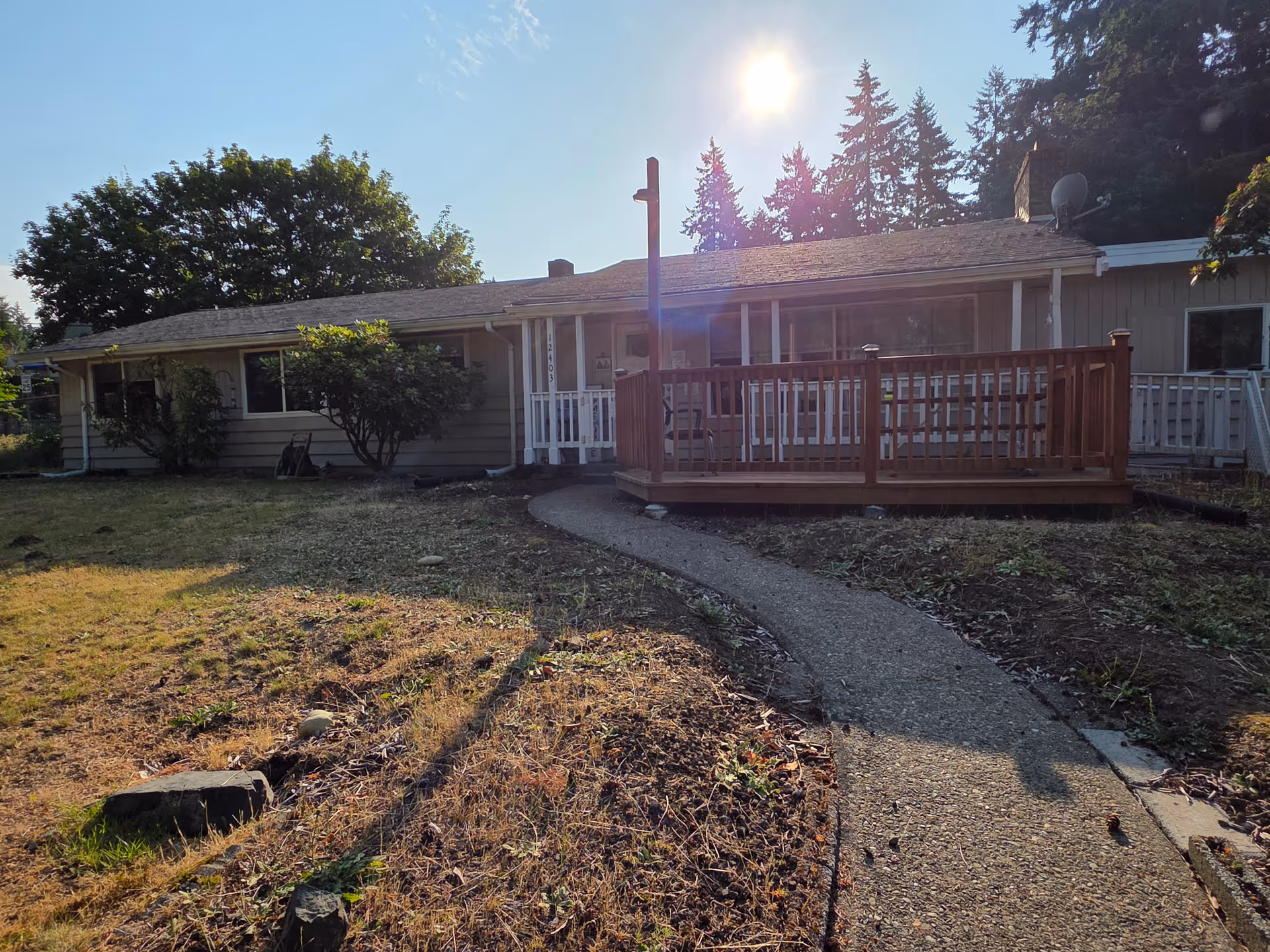 Single-story residential building with a wooden deck and a concrete pathway leading to the entrance, surrounded by dry grass and trees under a clear sky.