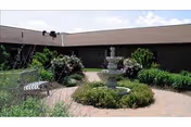 Outdoor courtyard area with a central stone fountain surrounded by greenery and flowering bushes, enclosed by a single-story building with a brown roof under a partly cloudy sky.
