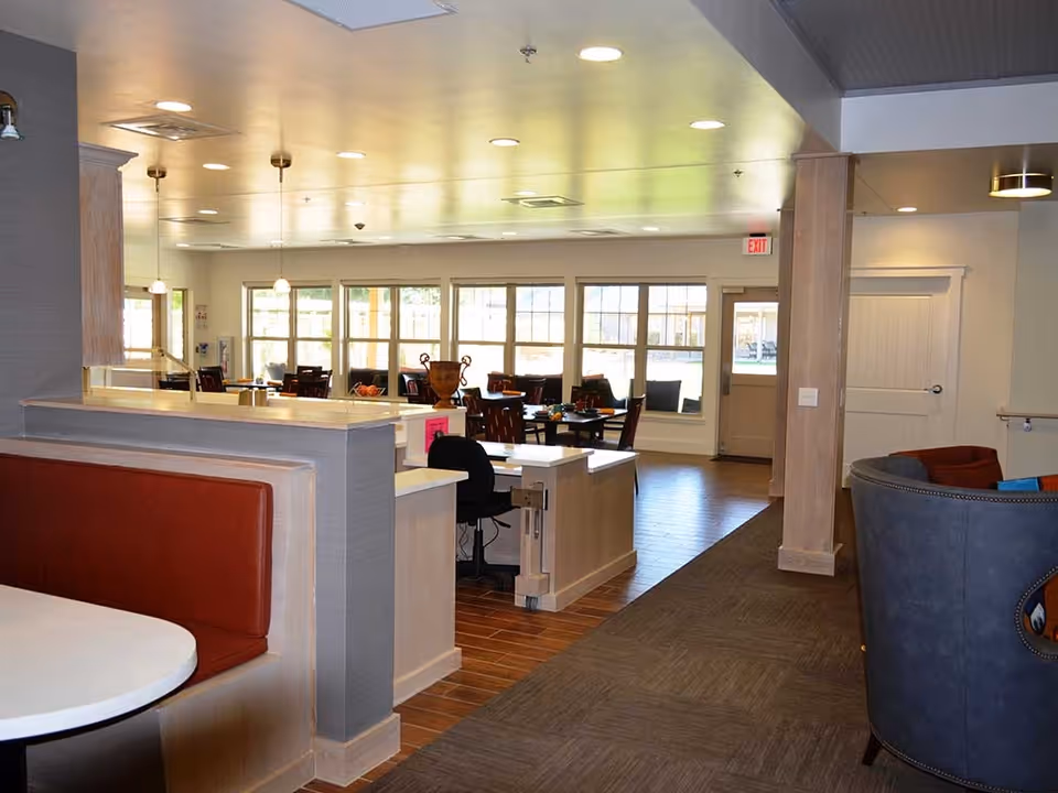 Interior view of a well-lit dining area in a senior living facility with multiple tables and chairs arranged near large windows. There is a booth seating area with red cushions in the foreground, a reception or service desk with a black office chair, and a lounge area with blue and red cushioned chairs on the right side. The floor is a combination of wood and carpet, and the ceiling has recessed lighting and pendant lights.