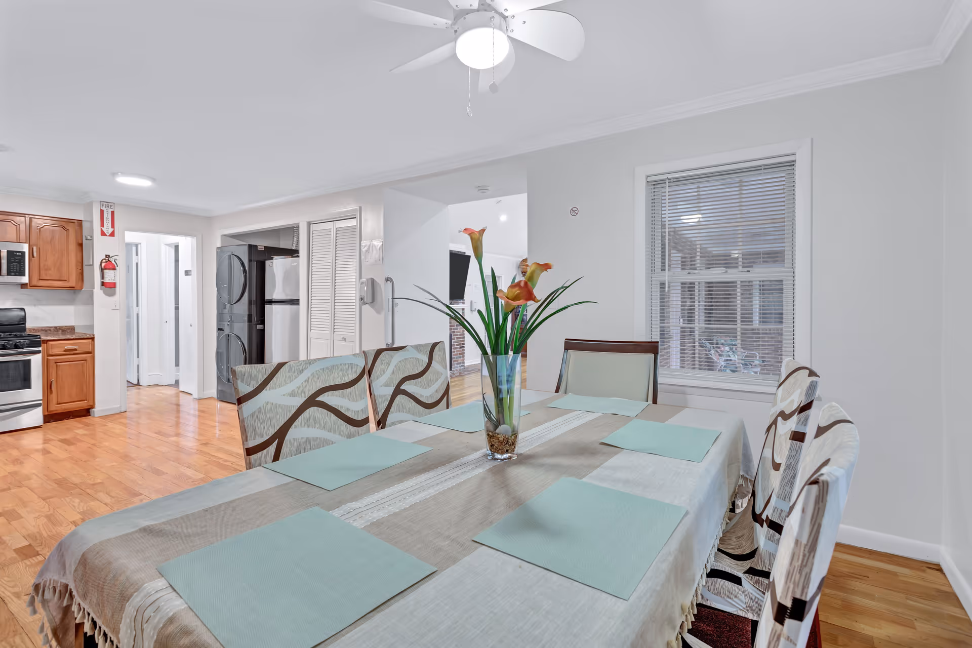 Bright dining room with a table set with placemats and a vase of flowers, adjacent to an open kitchen with wooden cabinets and stainless steel appliances.