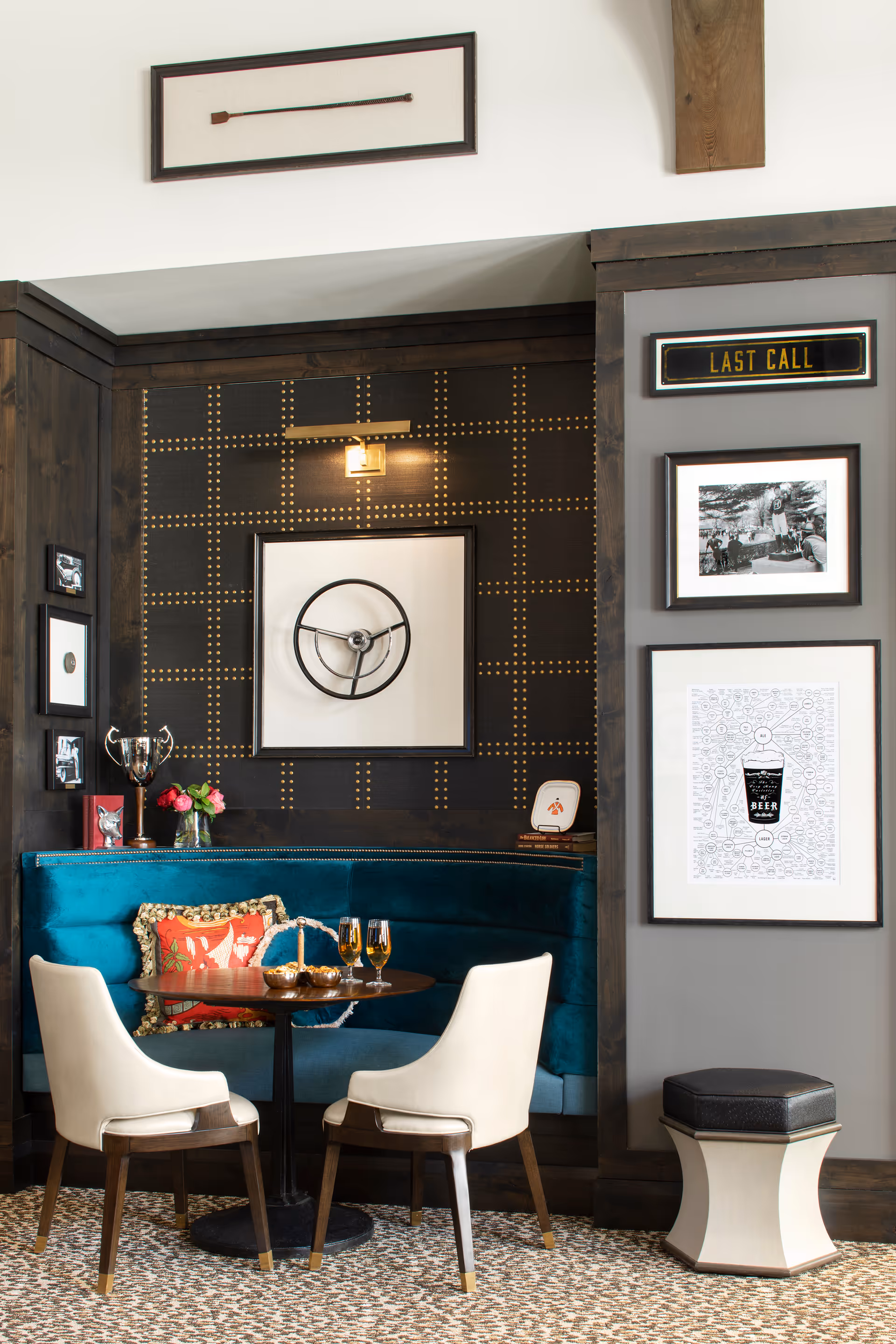 A cozy seating area with a round wooden table, two white cushioned chairs, and a teal velvet banquette with decorative pillows. The wall behind the banquette features a black and gold grid pattern with framed artwork, including a steering wheel and a 'LAST CALL' sign. There is a small stool to the right and a carpeted floor.