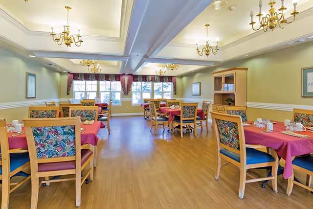 A bright dining room with multiple wooden tables covered with red tablecloths and surrounded by wooden chairs with floral patterned cushions. The room has large windows with burgundy valances, light green walls, wooden flooring, and several brass chandeliers hanging from a white ceiling with recessed lighting.