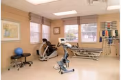 A physical therapy or rehabilitation room with exercise equipment including a stationary bike, a recumbent bike, a treatment table, a blue exercise ball, and medical supplies mounted on the wall. The room has large windows with blinds and framed artwork on the walls.
