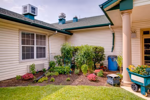 A small garden area with various green plants and flowers situated in the corner of a building with beige siding and green roof trim. There is a blue rain barrel, a green wheelbarrow with flowers, and a decorative stone planter near a column supporting a roof overhang.