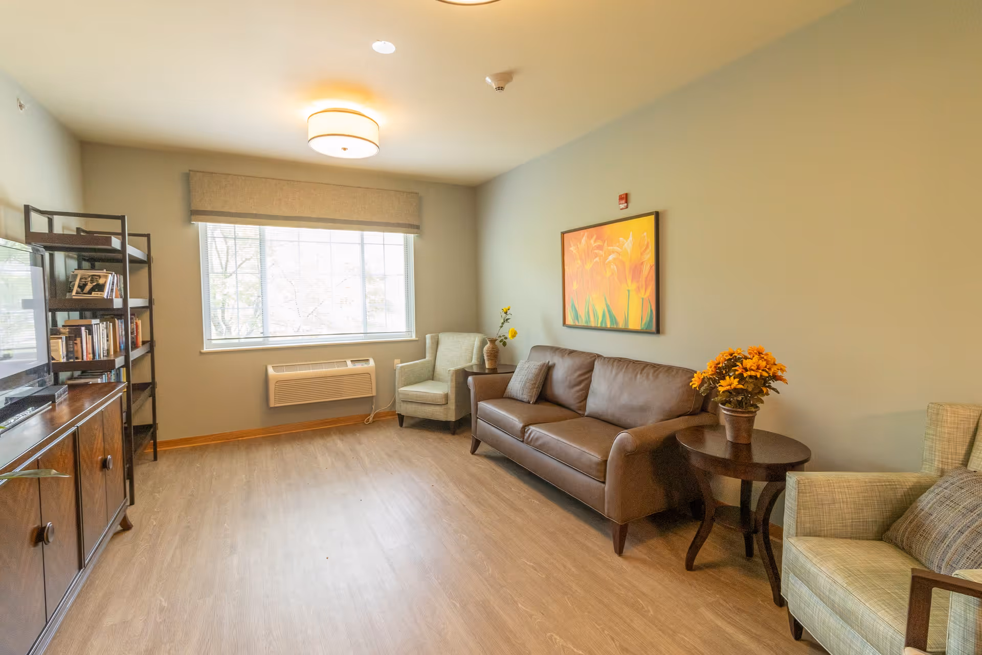 A cozy living room with a brown leather sofa, two armchairs, a wooden side table with a potted plant, a wooden TV stand with a television and bookshelf, a large window with a beige valance, and a colorful floral painting on the wall.