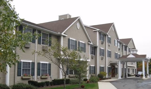 Exterior view of a multi-story senior living facility building with beige siding, dark green shutters, and a covered entrance with white columns. There are small trees and shrubs planted along the front of the building and a paved driveway leading to the entrance.
