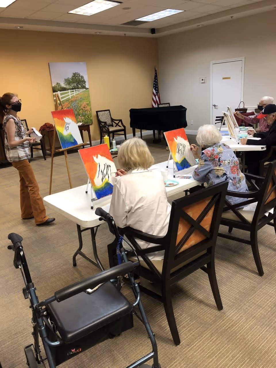 A group of elderly individuals seated at tables in a room, participating in a painting activity. Each person has a canvas with a colorful painting of a zebra on a red, yellow, and blue background. A woman wearing a mask stands nearby, possibly instructing or assisting the group. A walker is visible in the foreground, and an American flag and piano are in the background.