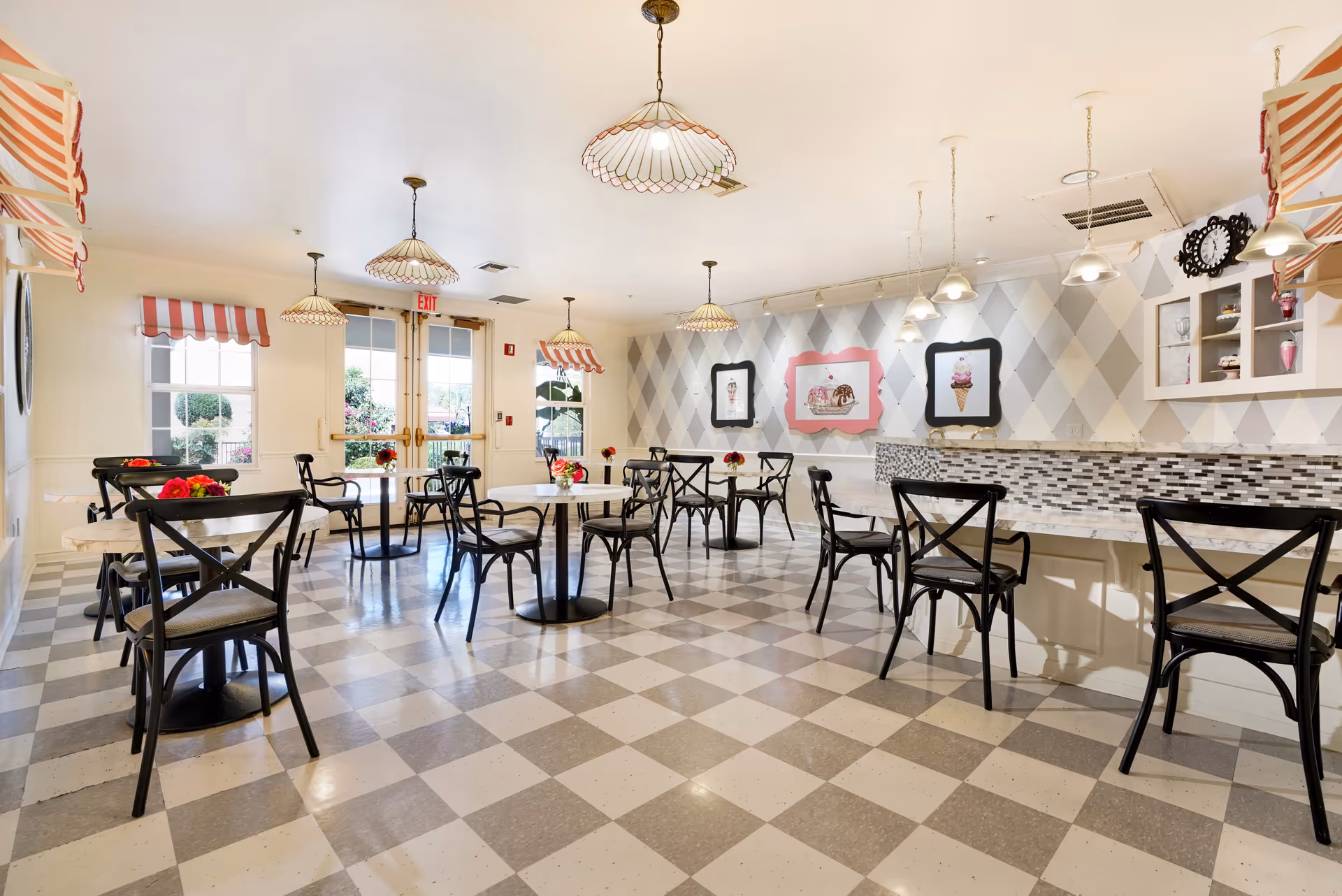 A bright and cheerful dining room with checkered floor tiles and several black chairs around white tables. The walls are decorated with framed pictures of desserts and an ice cream cone. There are pendant lights hanging from the ceiling and a counter with mosaic tile backsplash along one side. Windows and glass doors allow natural light to enter the room.
