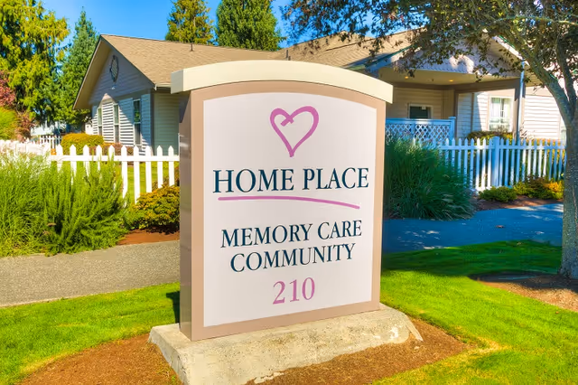 Freestanding sign in front of a picket-fenced home that reads "Home Place Memory Care Community 210" with landscaping and the facility building in the background.