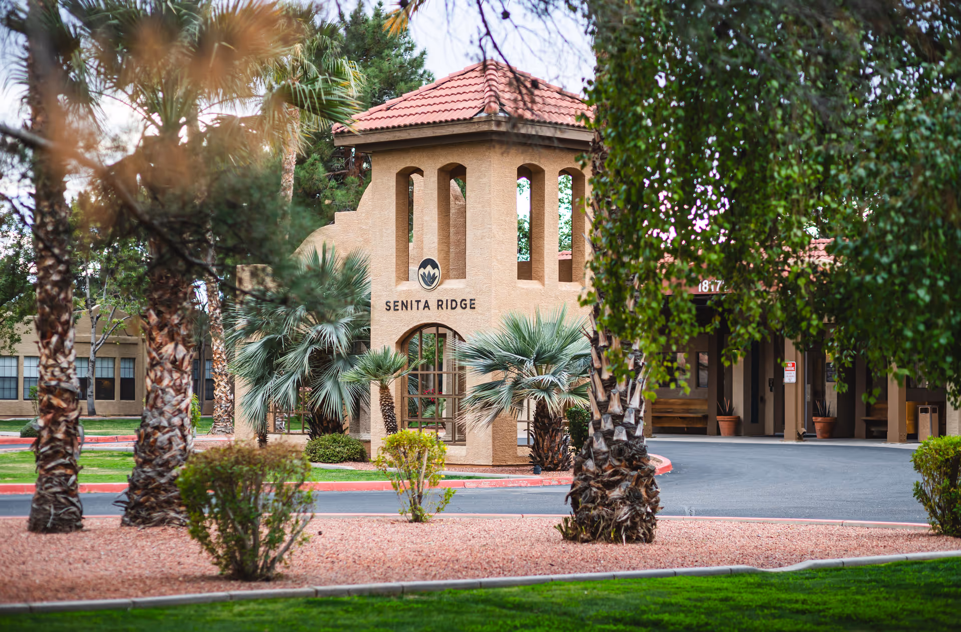 Exterior view of a senior living facility entrance with a beige stucco tower structure featuring arched windows and a red tile roof. The tower has a sign that reads 'SENITA RIDGE'. Surrounding the entrance are palm trees, bushes, and landscaped areas with red gravel and green grass. A driveway curves around the entrance, and part of the building is visible in the background.