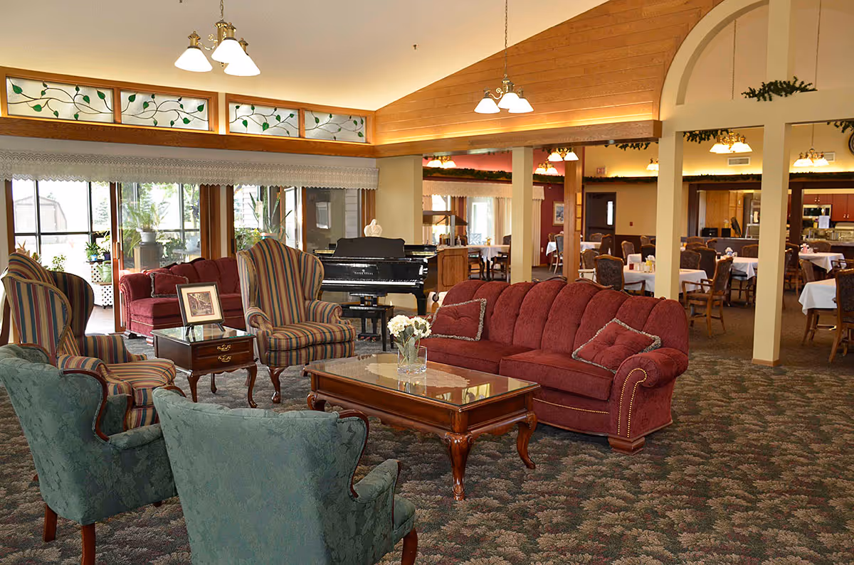 A cozy living room area in a senior living facility with a red sofa, two striped armchairs, two green armchairs, and a wooden coffee table with a vase of flowers. In the background, there is a black grand piano and a dining area with tables and chairs. The room has large windows with lace curtains and decorative stained glass panels near the ceiling.