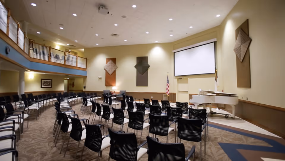 A large meeting or event room with rows of black chairs arranged facing a white projection screen and a white grand piano. The room has high ceilings, beige walls with decorative wall hangings, an American flag, and a balcony with wooden railings overlooking the space.