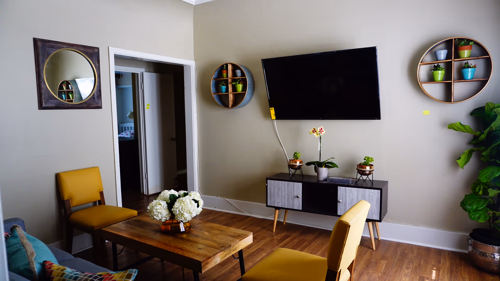 A cozy living room with wooden flooring, two yellow chairs, a wooden coffee table with a vase of white flowers, a wall-mounted flat screen TV above a modern TV stand with small potted plants, round wall shelves with colorful pots, a large green plant in a silver pot, and a round mirror on the wall reflecting part of the room.