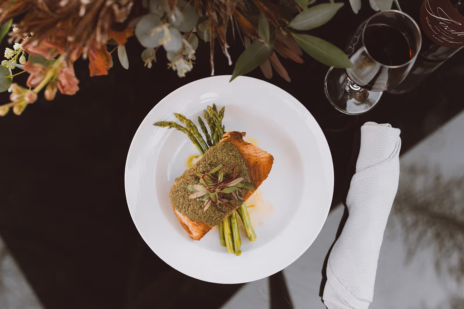A white plate with a serving of cooked salmon topped with green herb crust and garnished with microgreens, placed on a bed of asparagus spears. Next to the plate is a neatly folded white napkin and a glass of red wine on a dark reflective surface, with some floral decorations partially visible at the top left corner.