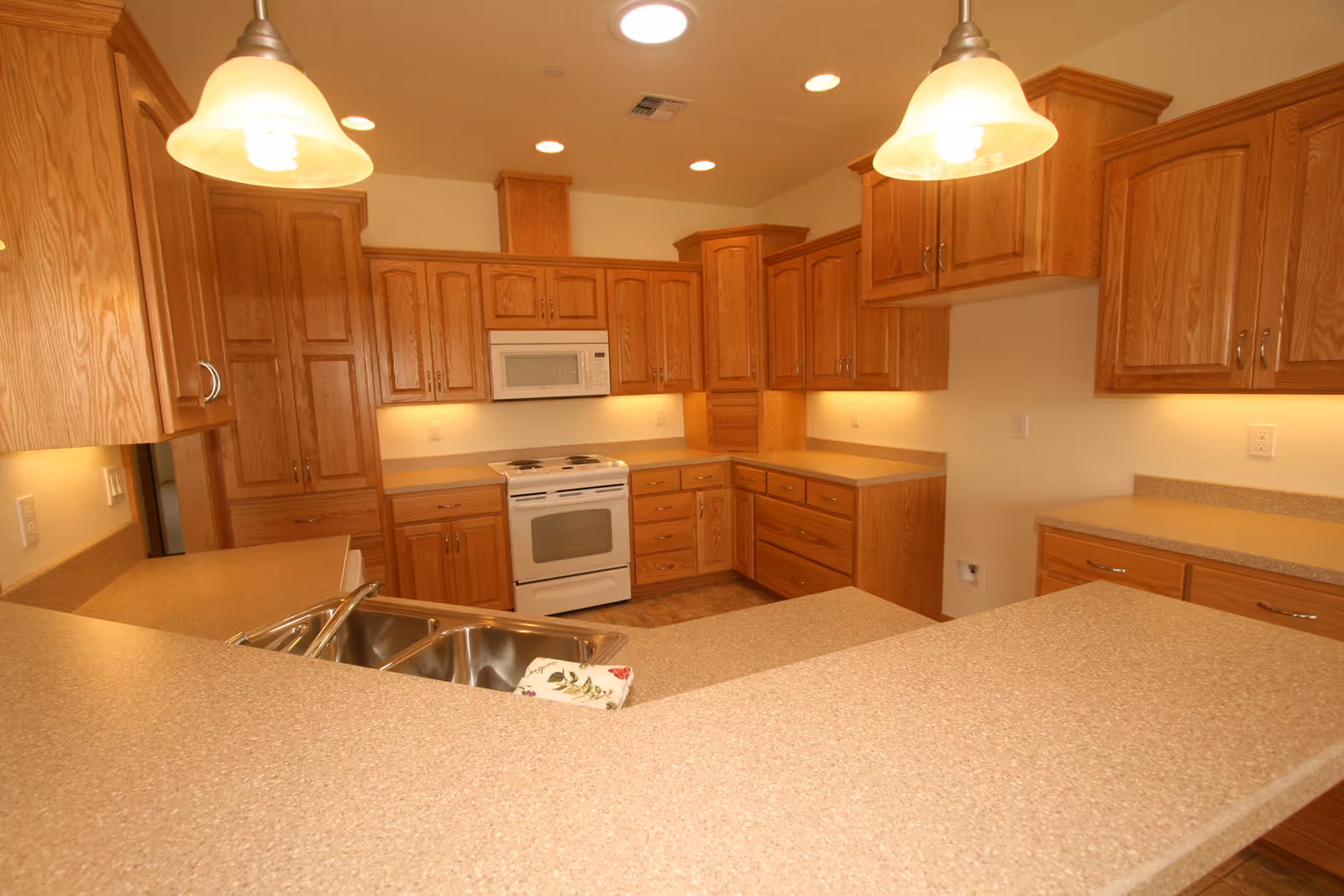 A spacious kitchen with wooden cabinets, beige countertops, a double sink, a white stove, and a microwave. Two pendant lights hang from the ceiling, illuminating the area.