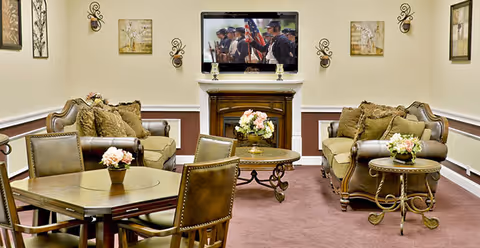 Cozy assisted-living common room with sofas and armchairs arranged around a coffee table, a TV above a fireplace, and floral centerpieces.
