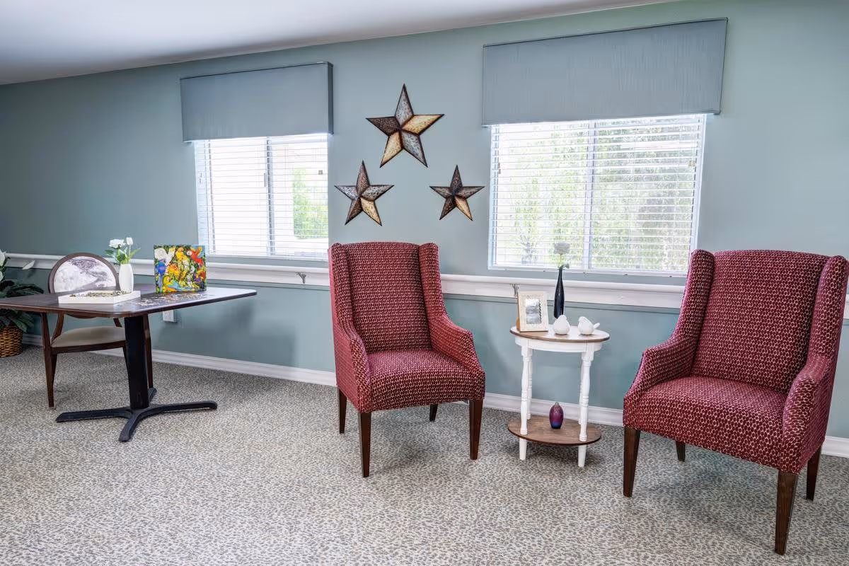 A cozy interior room with two red patterned armchairs separated by a small round white table holding decorative items. Behind the chairs are two windows with blinds and gray valances. On the wall between the windows are three decorative star-shaped wall hangings. To the left, there is a table with a chair and some decorative items on it, and a plant in the corner.