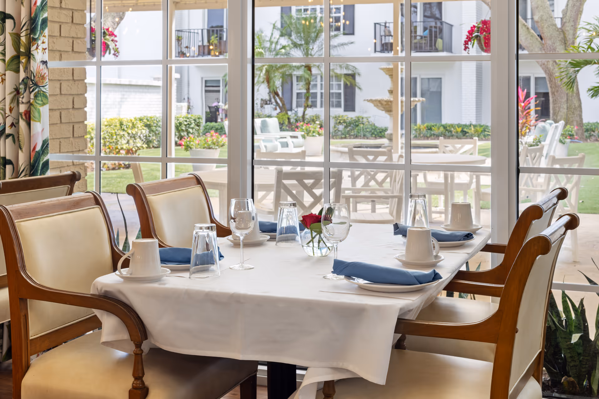 A dining table set for four with white tablecloth, blue napkins, white cups, saucers, and wine glasses in a senior living facility. The table is positioned near large windows that overlook a garden with outdoor seating, plants, and a fountain.