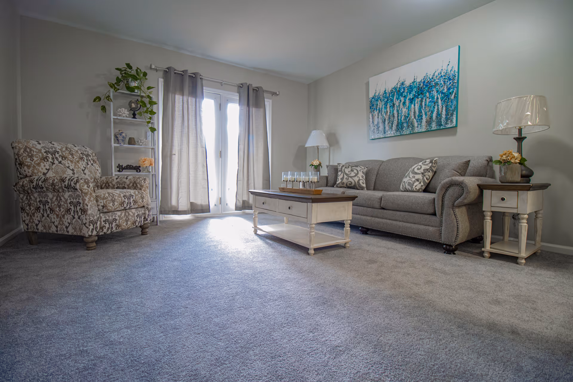 Spacious living room with a gray sofa, patterned armchair, coffee table, and large window with curtains.