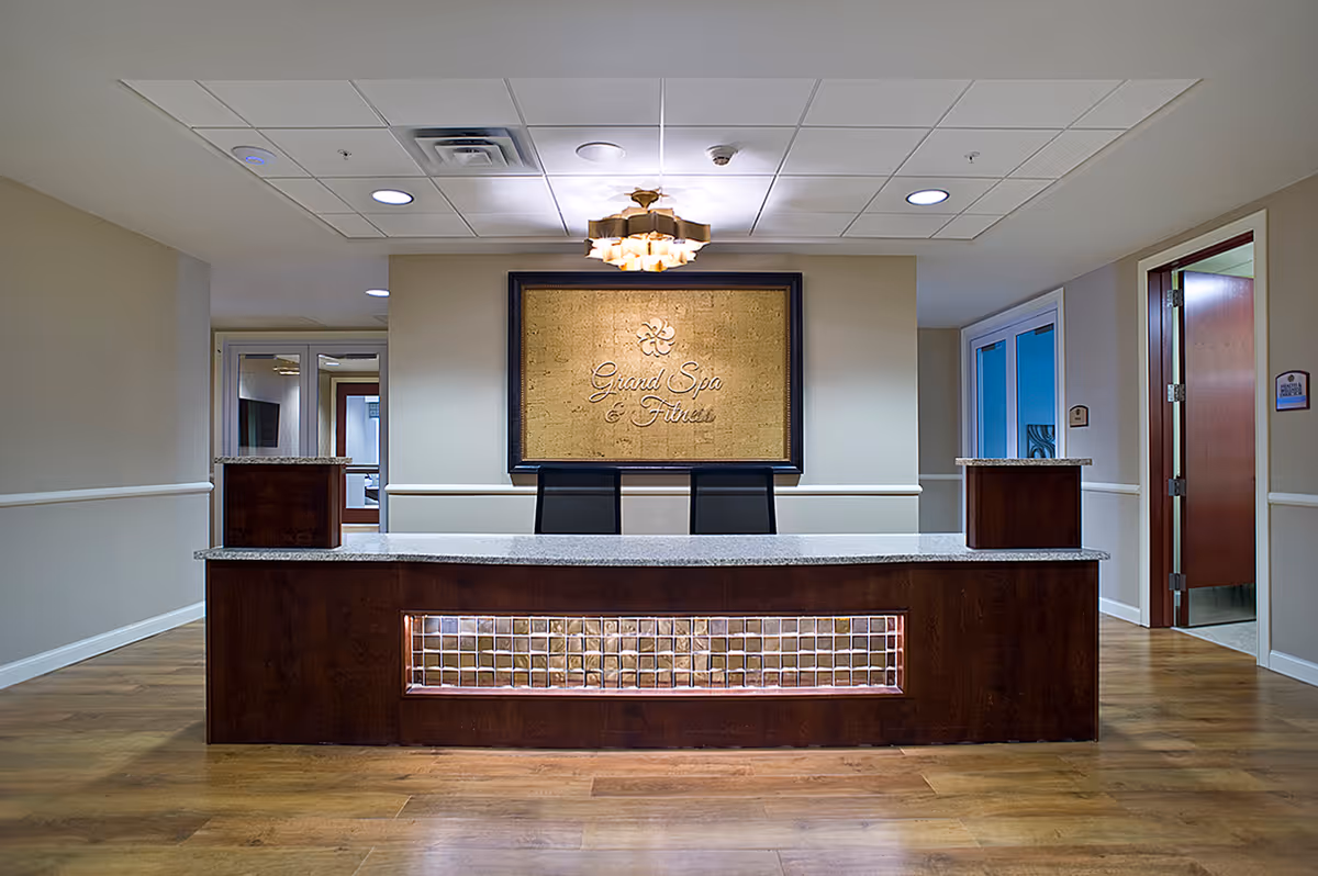 Reception desk area with a granite countertop and dark wood base featuring glass block accents. Behind the desk is a framed sign that reads 'Grand Spa & Fitness' mounted on a beige wall. The floor is wood, and the ceiling has recessed lighting and a decorative chandelier. Doors and hallways are visible on either side of the desk.