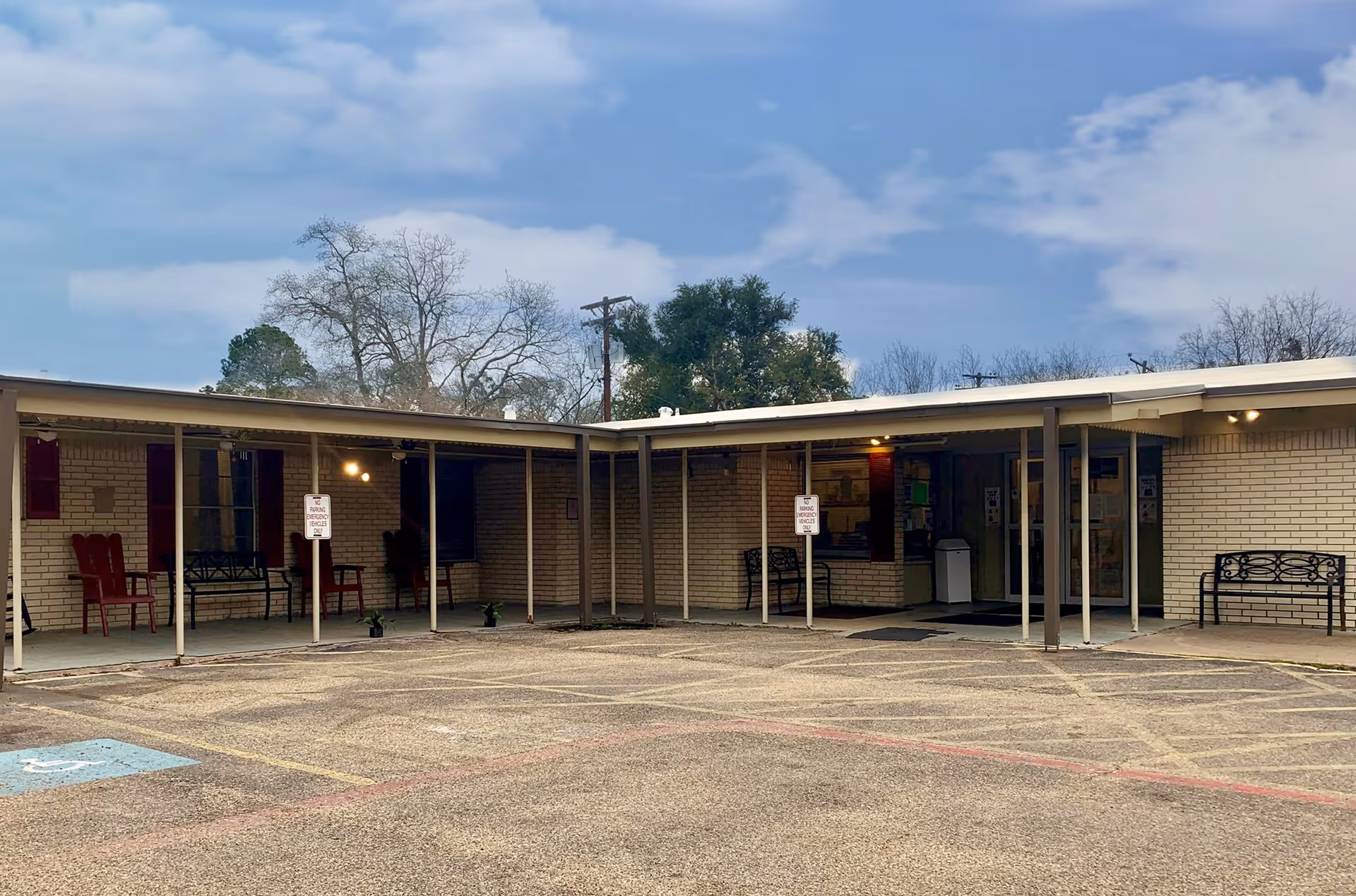 Exterior view of a single-story brick building with a covered walkway supported by metal poles. Several benches and chairs are placed under the walkway. The sky is partly cloudy and there are trees in the background. The parking area in front has marked spaces including a handicapped spot.