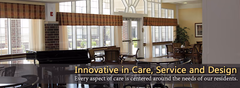 Interior view of a senior living facility common area with large windows, multiple round tables and chairs, a piano, and a door leading outside. The space is well-lit with natural light and decorated with plants and framed artwork.