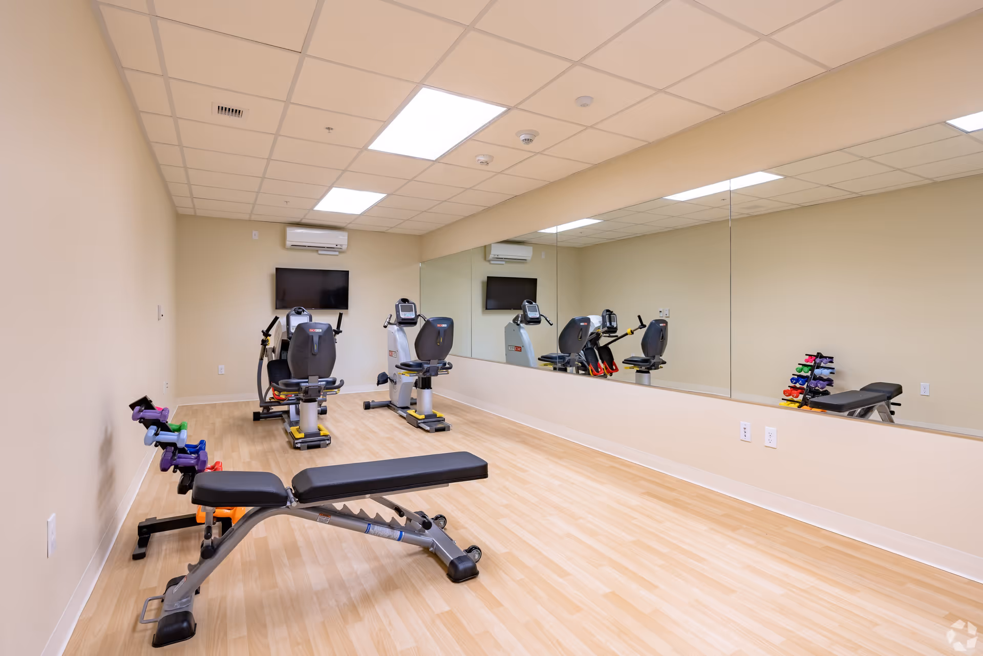 Small exercise room with mirrors, two recumbent bikes, a workout bench and a rack of colorful dumbbells.